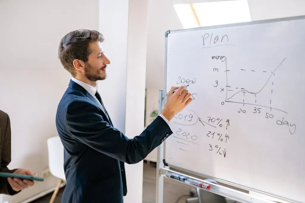 A businessman in a suit writes financial data on a whiteboard during an office planning session.